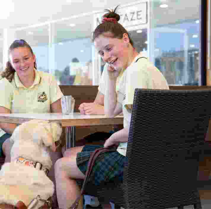 Two young adults sitting a cafe table. A yellow Guide Dog in harness is seated next to the table looking up at the people. The young adults are looking down at the Guide Dog smiling.