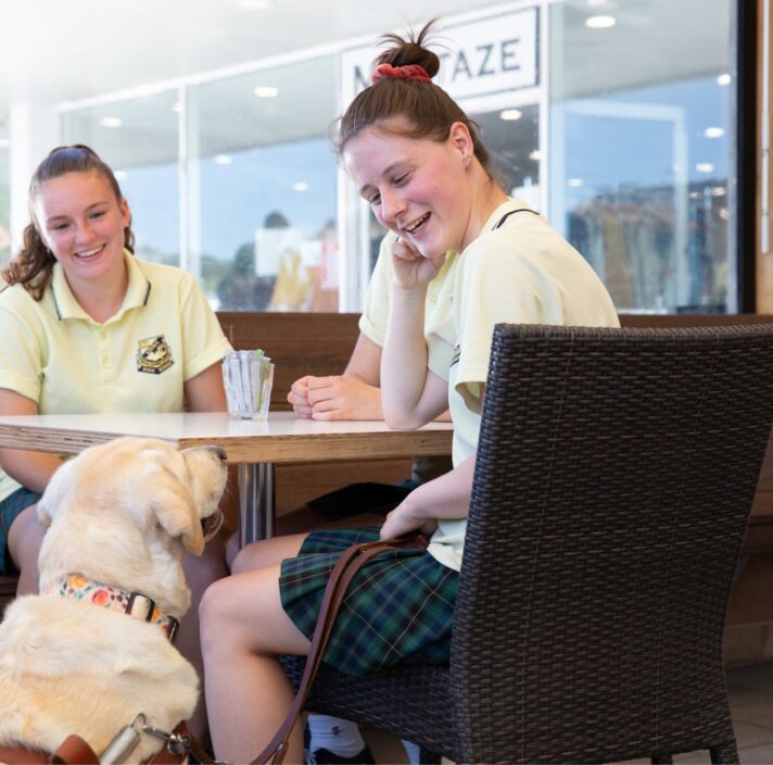 Two young adults sitting a cafe table. A yellow Guide Dog in harness is seated next to the table looking up at the people. The young adults are looking down at the Guide Dog smiling.