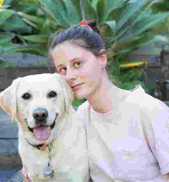 A person looking straight at the camera with their arm around a yellow labrador dog. They are seated at the bottom of a set of stairs.