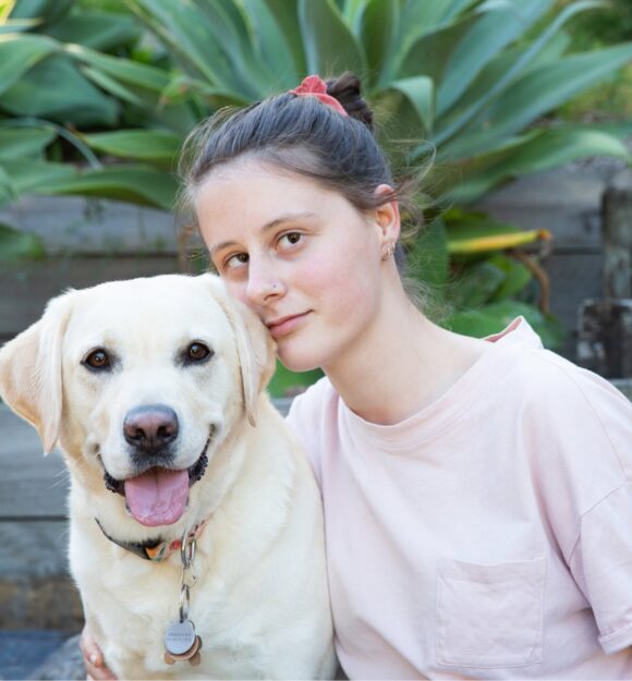 A person looking straight at the camera with their arm around a yellow labrador dog. They are seated at the bottom of a set of stairs.