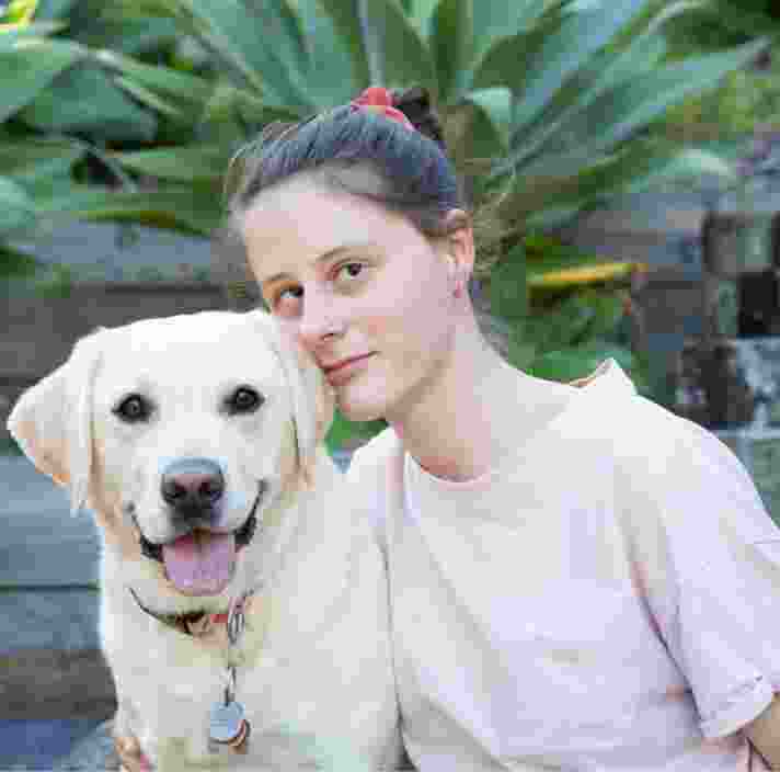A person looking straight at the camera with their arm around a yellow labrador dog. They are seated at the bottom of a set of stairs.