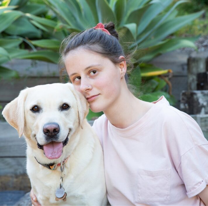 A person looking straight at the camera with their arm around a yellow labrador dog. They are seated at the bottom of a set of stairs.