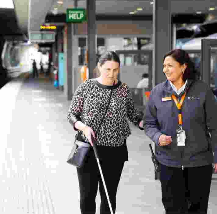 A person using their white cane at the train station while being guided by a train station attendant.