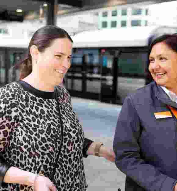 A person using their white cane at the train station while being guided by a train station attendant.