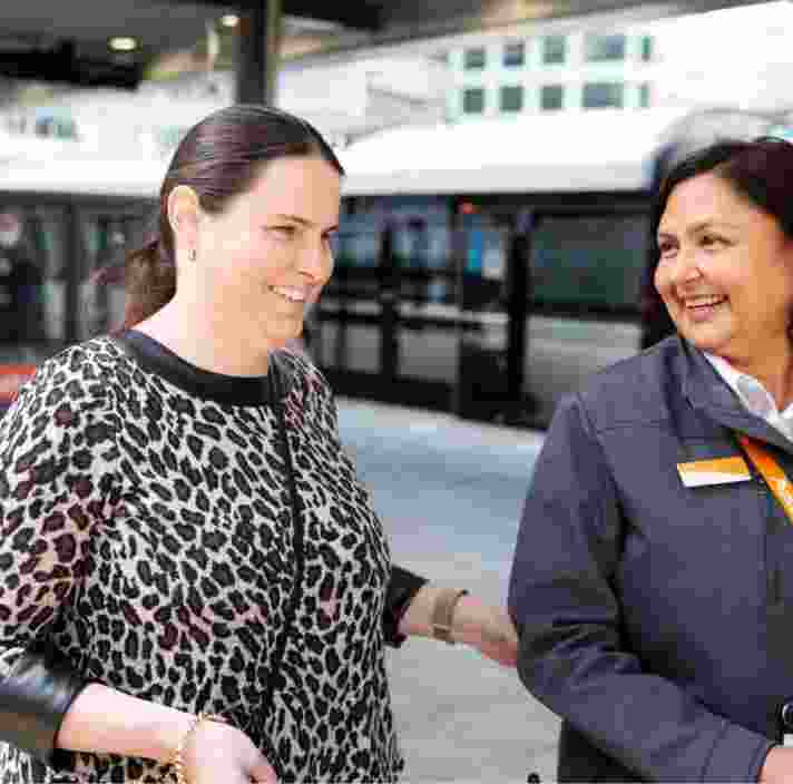 A person using their white cane at the train station while being guided by a train station attendant.