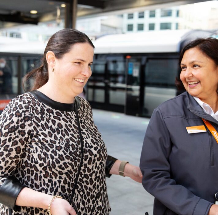 A person using their white cane at the train station while being guided by a train station attendant.
