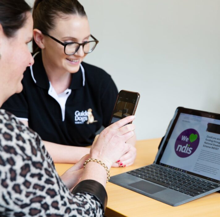 A person and a Guide Dog's staff member looking at a laptop. The person is holding up an Iphone to the laptop and the staff member is smiling.