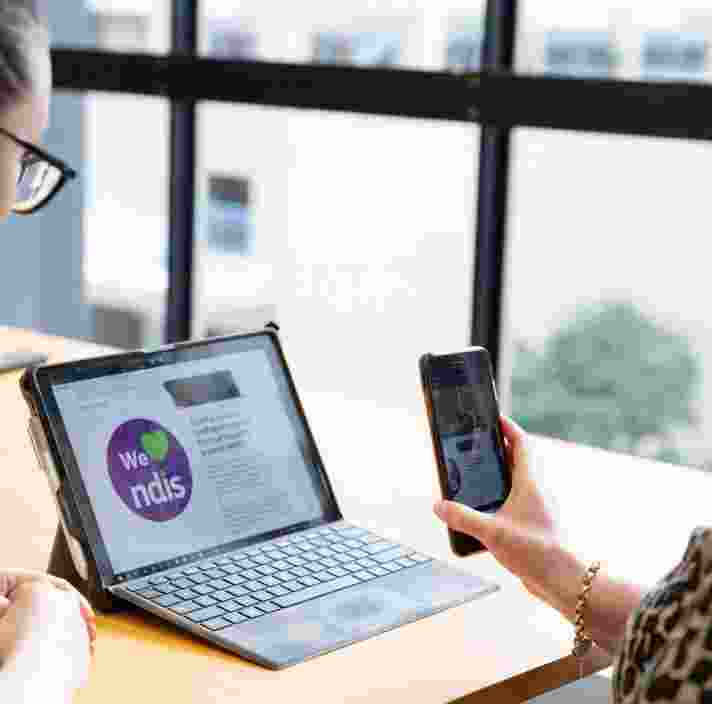 Two people looking at a laptop screen. One is holding a phone to the laptop screen and the other is watching on.