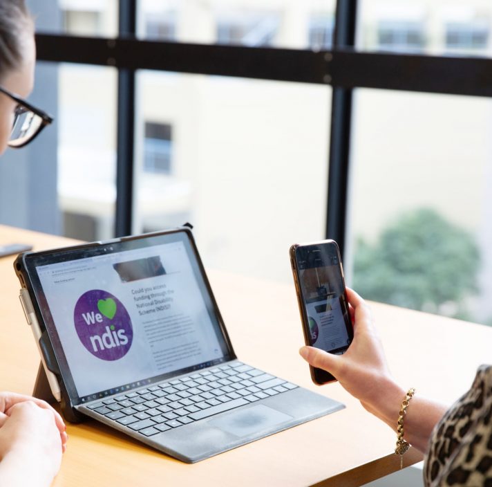 Two people looking at a laptop screen. One is holding a phone to the laptop screen and the other is watching on.