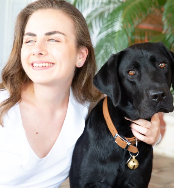 A person looking to the left of the camera smiling with their arm around the neck of a black labrador dog.