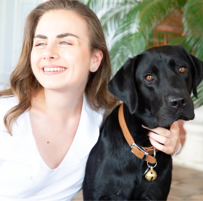 A person looking to the left of the camera smiling with their arm around the neck of a black labrador dog.