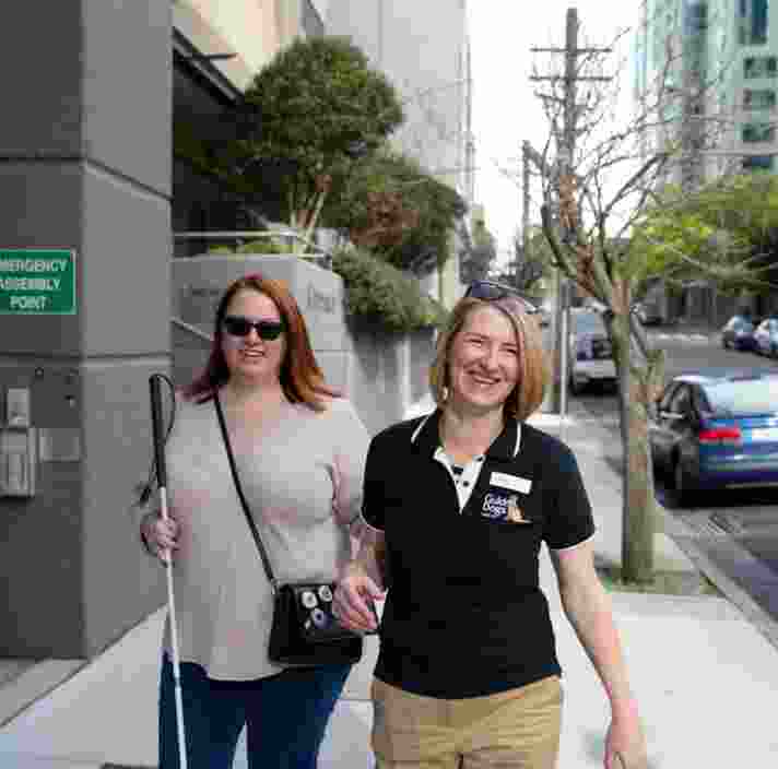 A person with their white cane walking down the street with a Guide Dogs staff member. Both people are smiling at the camera and the person with the cane is holding the staff member by the elbow.
