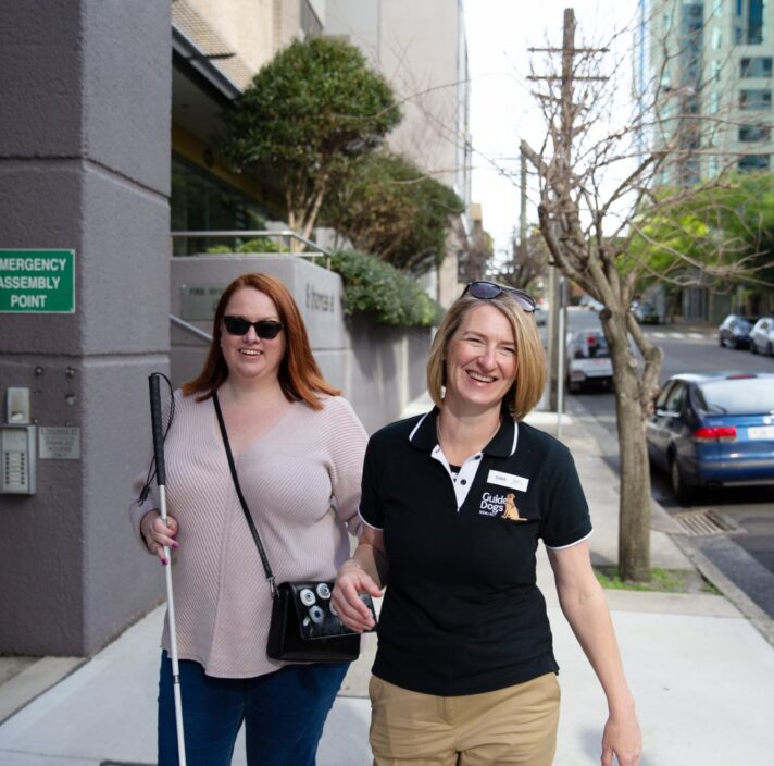 A person with their white cane walking down the street with a Guide Dogs staff member. Both people are smiling at the camera and the person with the cane is holding the staff member by the elbow.