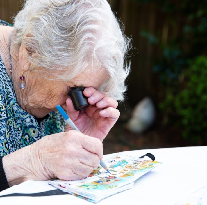 An older adult using a magnifier whilst painting, The person is painting onto a canvas.