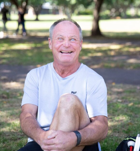 A person sitting outside on the grass with a smile on their face looking at the camera. They have one leg bent with their arm wrapped around their knee and a white cane next to them.
