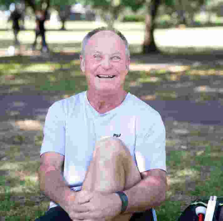A person sitting outside on the grass with a smile on their face looking at the camera. They have one leg bent with their arm wrapped around their knee and a white cane next to them.