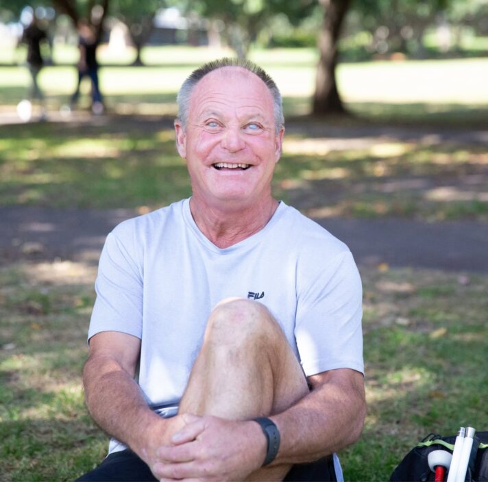 A person sitting outside on the grass with a smile on their face looking at the camera. They have one leg bent with their arm wrapped around their knee and a white cane next to them.