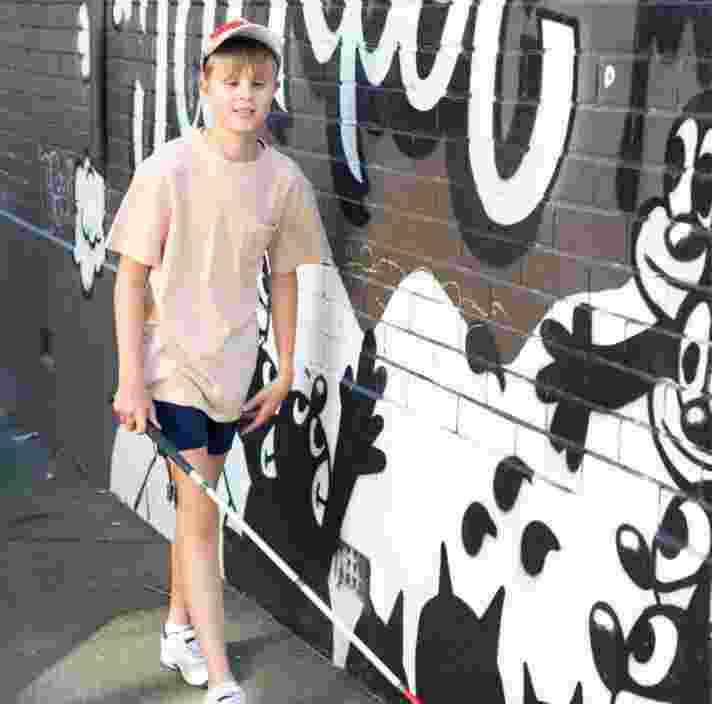 A child walking down the street using their white cane. The child is walking against a wall that has graffiti on it.
