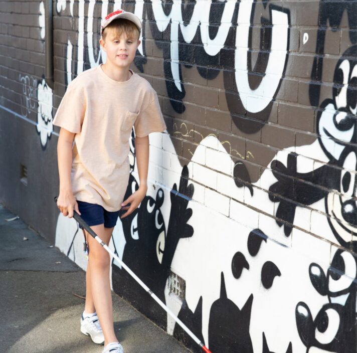 A child walking down the street using their white cane. The child is walking against a wall that has graffiti on it.