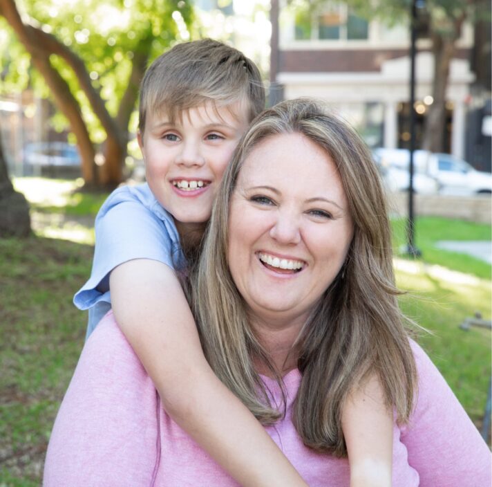 Young child with their arms around their mothers neck and they are both looking at the camera smiling. They are seated on grass outside.