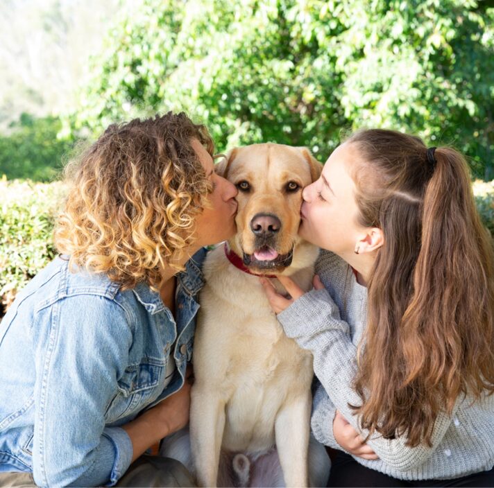 Two people kissing the cheeks of a yellow labrador dog. The dog is seated in between the two people and is looking at the camera.
