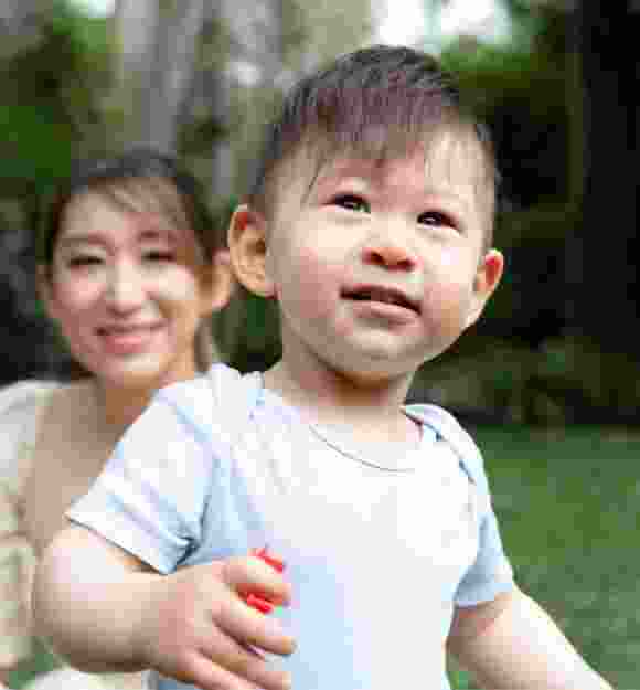 A young child playing outside with the mother watching in the background.