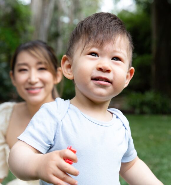 A young child playing outside with the mother watching in the background.