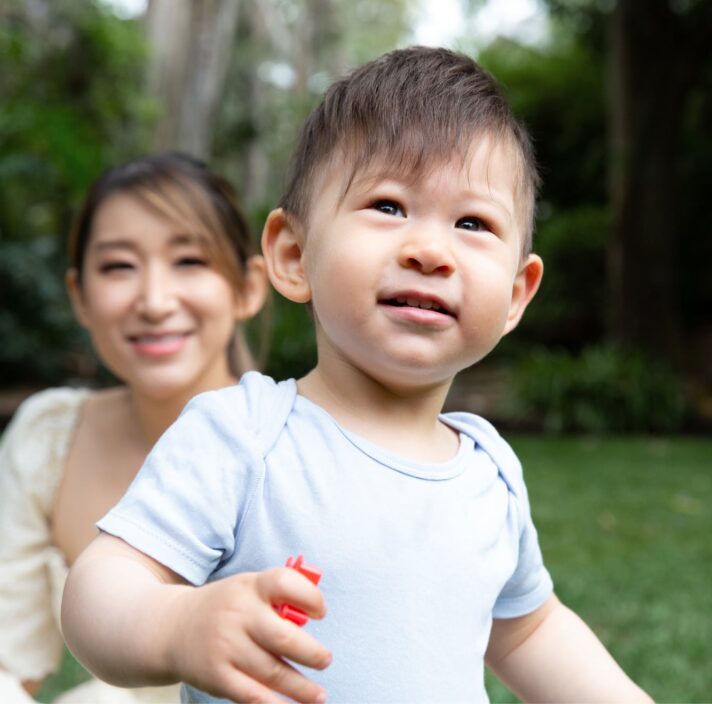A young child playing outside with the mother watching in the background.
