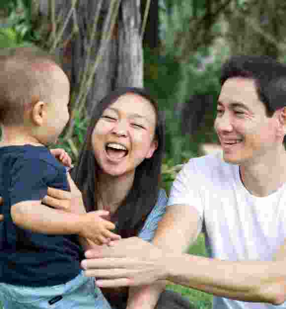 A couple sitting outside watching their baby walk towards them. The mother and father have smiles on their faces.
