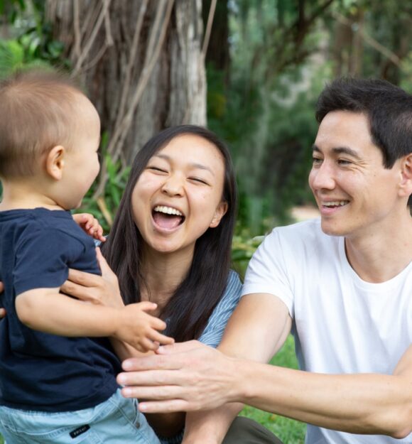 A couple sitting outside watching their baby walk towards them. The mother and father have smiles on their faces.