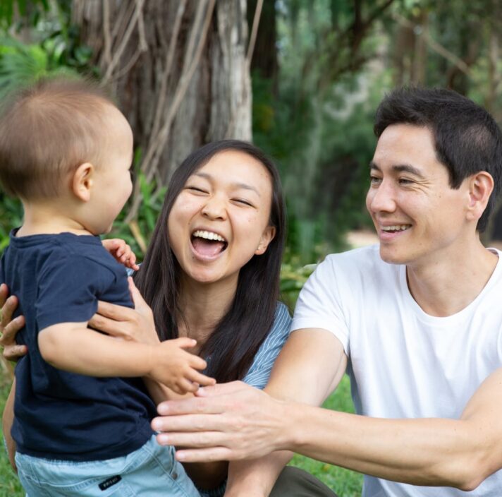 A couple sitting outside watching their baby walk towards them. The mother and father have smiles on their faces.