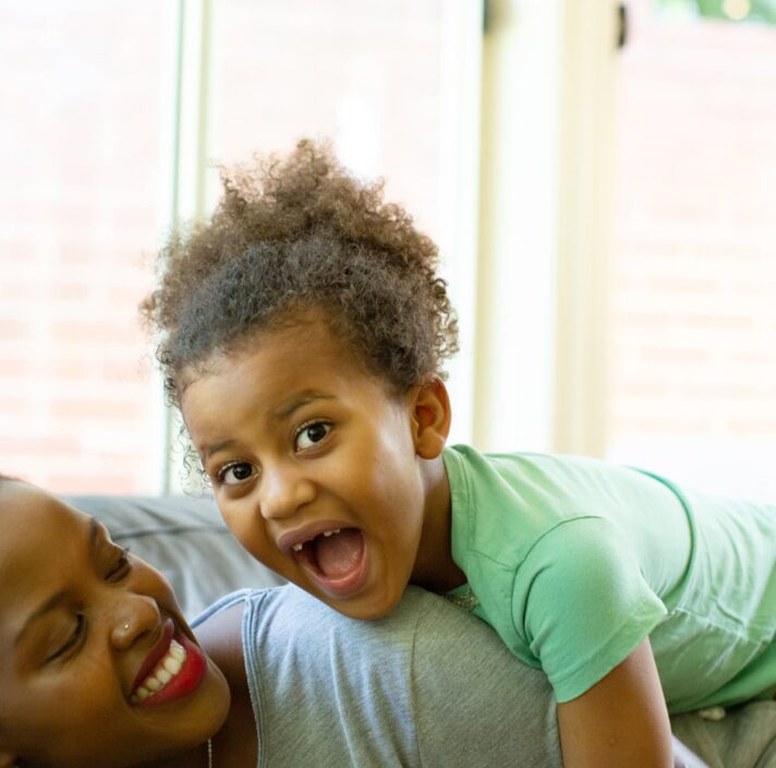A mother and young child playing on the couch. The mother the child on top of her and the child has a big smile on their face.