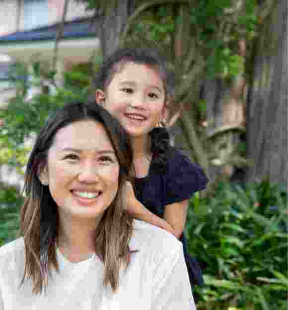 A mother and young child outside smiling at the camera. The young child is standing behind the mother with their hands on her back.
