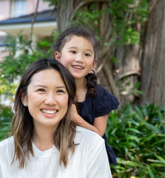 A mother and young child outside smiling at the camera. The young child is standing behind the mother with their hands on her back.