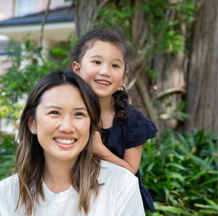 A mother and young child outside smiling at the camera. The young child is standing behind the mother with their hands on her back.