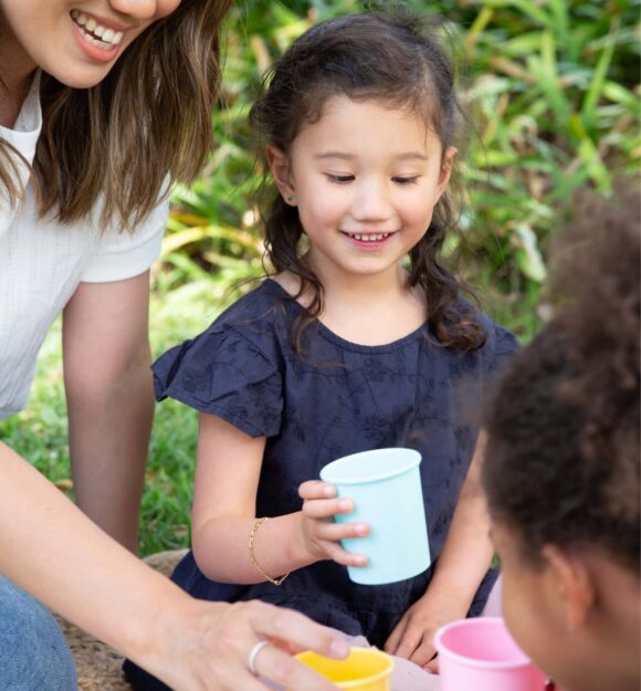 Two young children and a mother are playing with a tea set outside. The children and mother are smiling.
