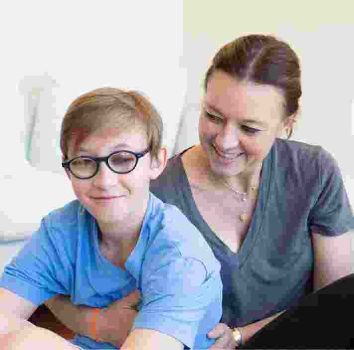A young child with their mother seated on the ground. The mother is holding the child from behind and looking at her child. The child is looking at the camera smiling.