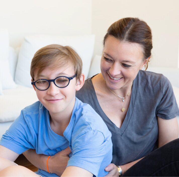 A young child with their mother seated on the ground. The mother is holding the child from behind and looking at her child. The child is looking at the camera smiling.