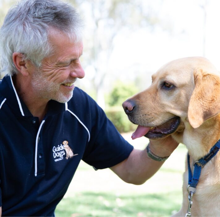 A Guide Dogs staff member and a yellow labrador dog sitting outside. The staff member is patting the dog whilst smiling and they are both looking at each other.