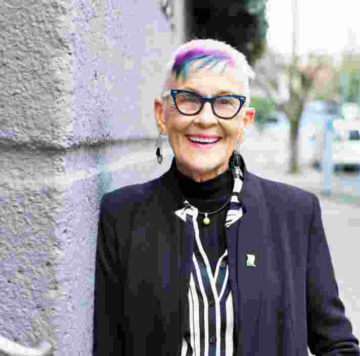 A person standing outside and leaning against a wall looking at the camera with a smile on their face.