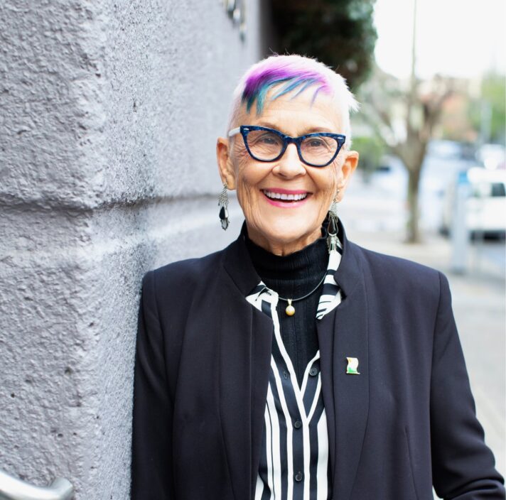 A person standing outside and leaning against a wall looking at the camera with a smile on their face.
