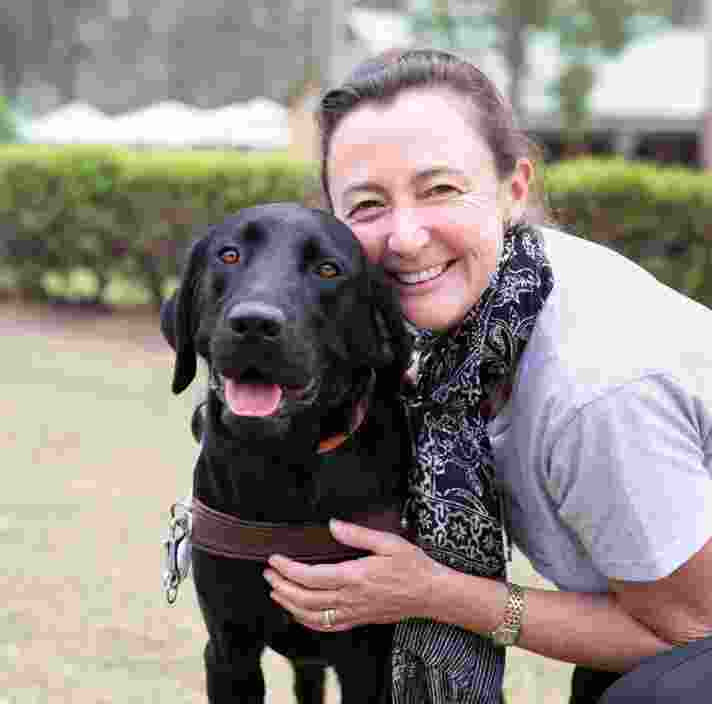 A person hugging a black labrador Guide Dog in harness. The person and dog are looking at the camera and the person is smiling,