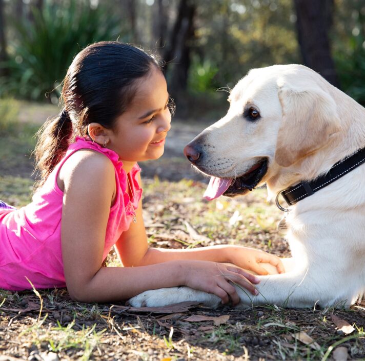 A young child laying down looking into the face of a yellow labrador dog with a smile on their face, They are both outside and are sitting facing each other.