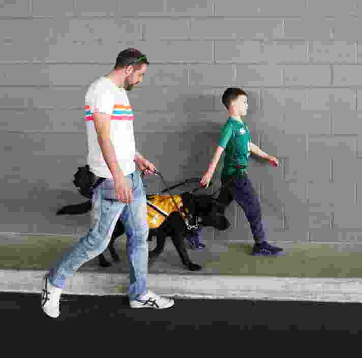 A man and a young child walking along a street. The child and man are holding the leash of a black labrador dog who is wearing an Autism Assistance Dog jacket.