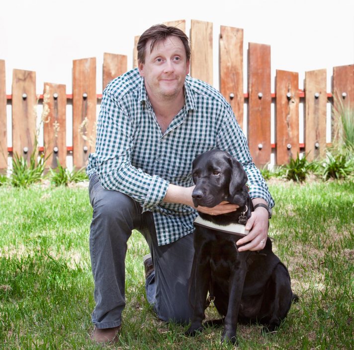 A person kneeling down outside next to a black labrador Guide Dog in harness.