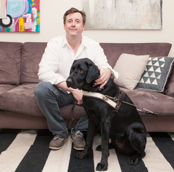 A person seated on a couch inside with a black labrador Guide Dog in harness next to them.