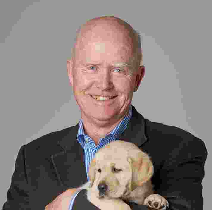 Headshot of Guide Dogs Victoria's Board Director, Bruce Brook. Bruce is looking at the camera smiling and holding a yellow eight week old labrador puppy.