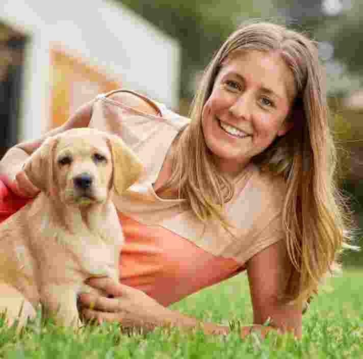A person laying outside on some grass next to an eight week old labrador puppy. The puppy and person are both looking at the camera.