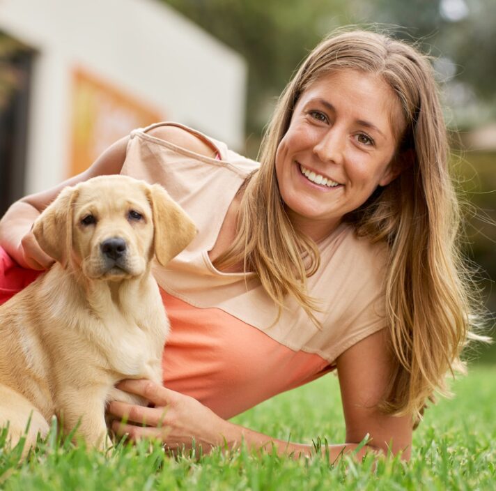 A person laying outside on some grass next to an eight week old labrador puppy. The puppy and person are both looking at the camera.