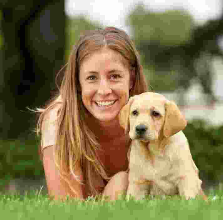 A person laying on some grass with an eight week old yellow labrador puppy. The person is smiling and looking at the camera. The puppy is also looking at the camera.